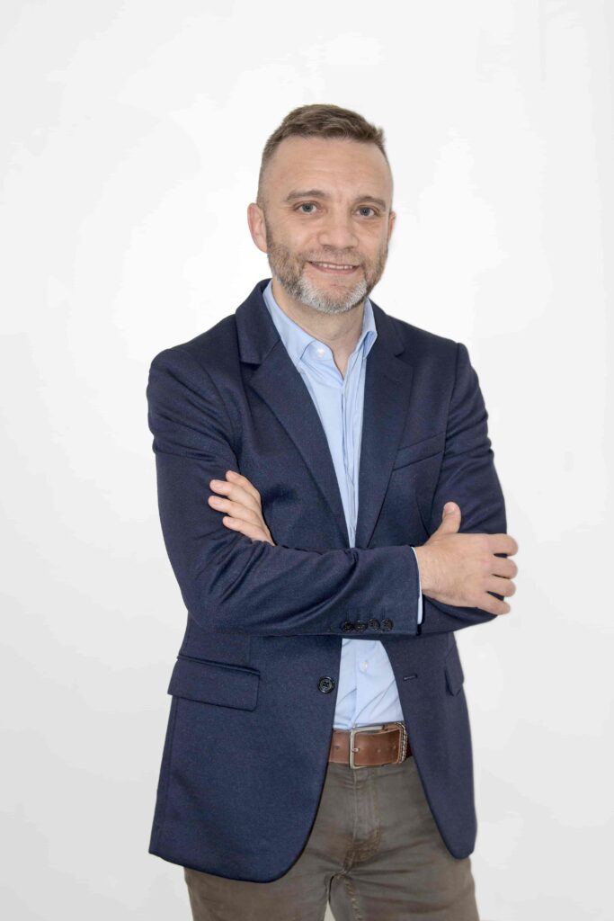 Hombre mediana edad con cabello corto y barba. Tiene ojos color azul. Posa sonriendo y cruzando los brazos. Lleva puesto una americana azul oscuro y una camisa azul clara.