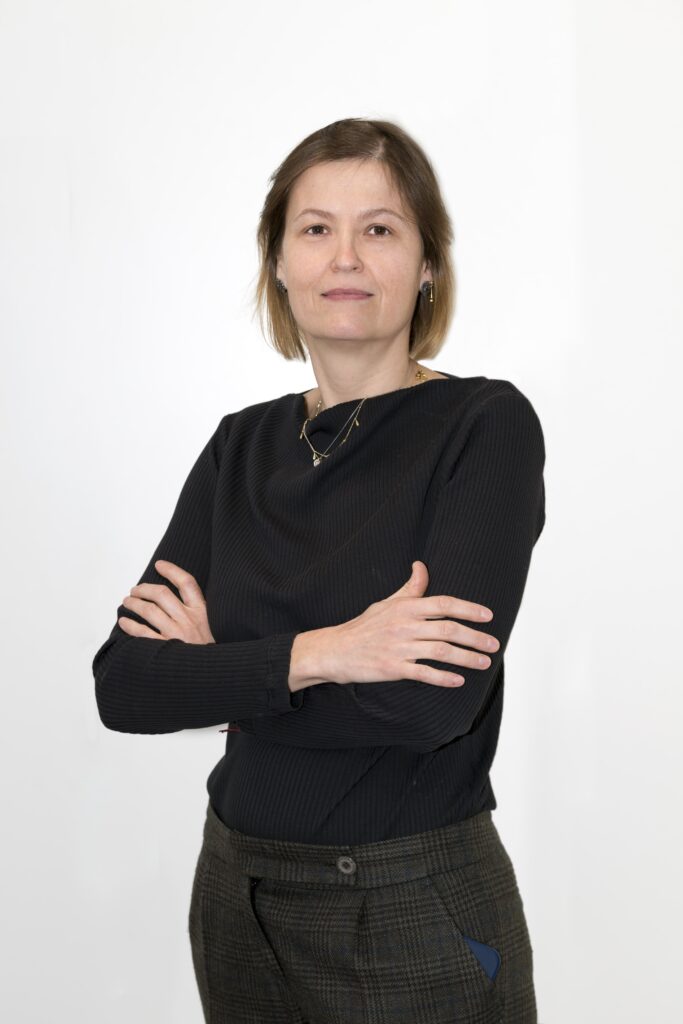 Mujer mediana edad con cabello corto por encima de los hombros de color castaño. Tiene ojos color marron oscuro. En la foto aparece con una ligera sonrisa sin mostrar los dientes y cruzando los brazos