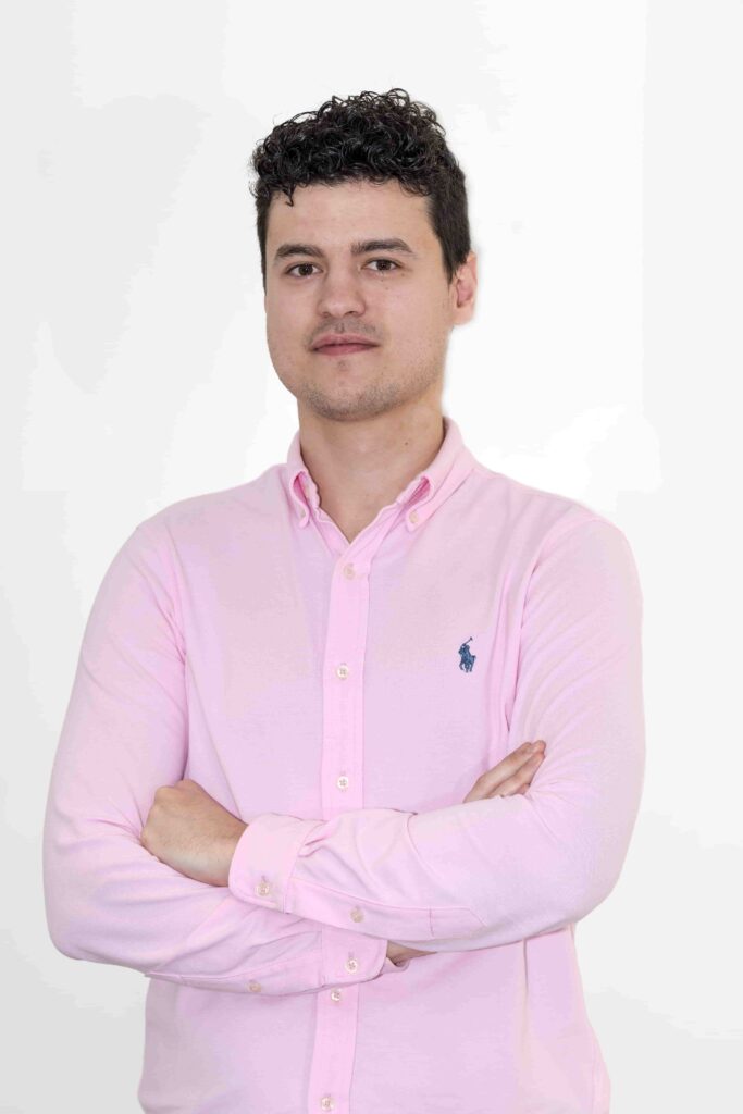 Hombre joven con cabello oscuro rizado. Tiene ojos color oscuro y en la foto aparece con una ligera sonrisa y con los brazos cruzados