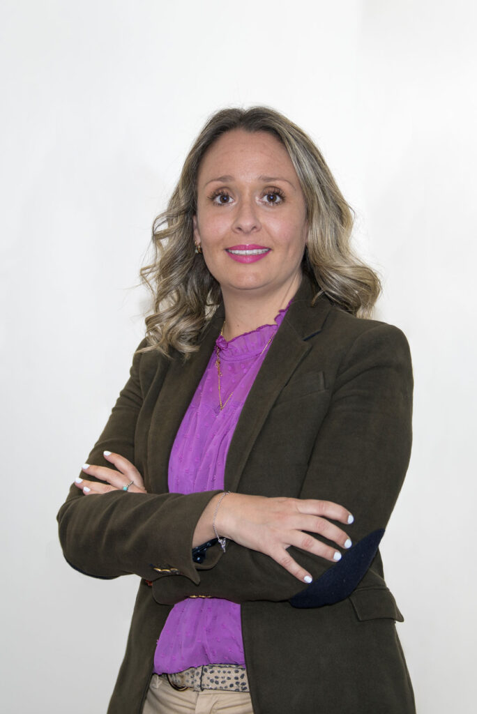 Mujer mediana edad con cabello rubio corto. Tiene ojos color marrón oscuro En la foto aparece con los brazos cruzados sonriendo