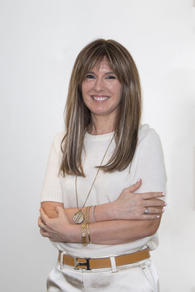 Mujer mediana edad con cabello largo de color castaño. Tiene ojos marrón oscuro. En la foto aparece con los brazos cruzados sonriendo